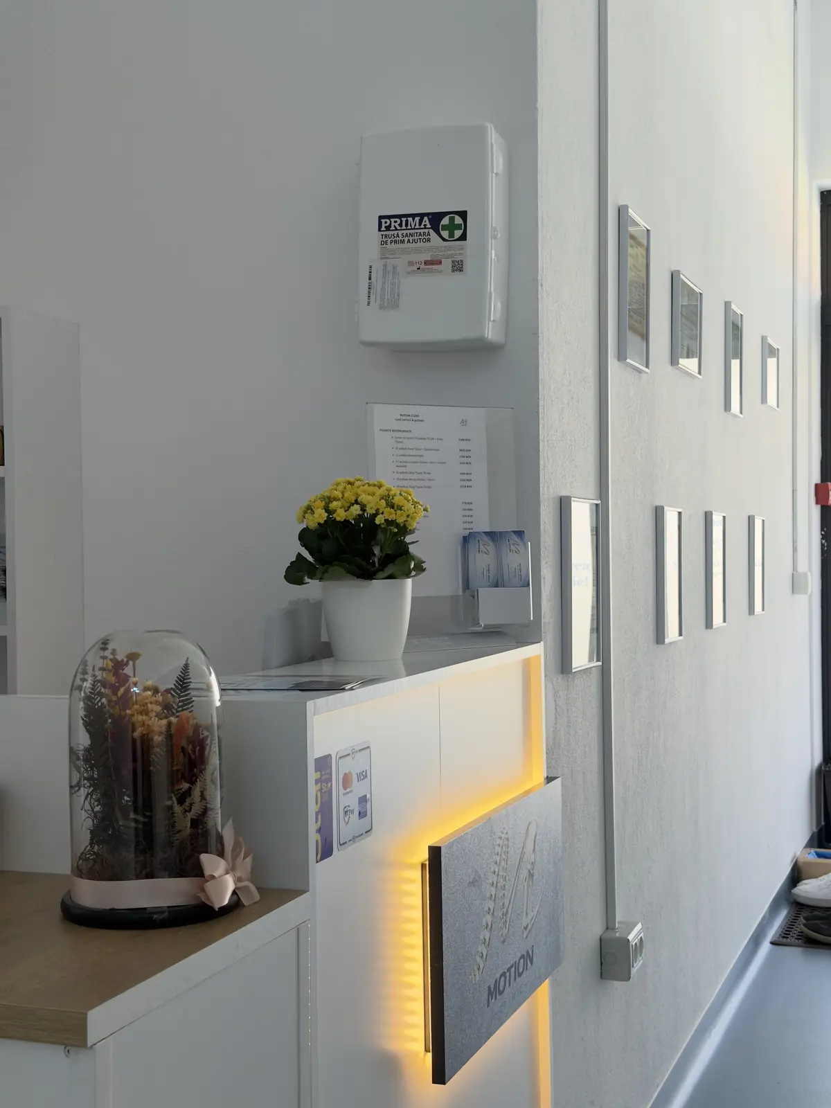 Recepție Motion Clinic: tejghea albă cu logo luminat, flori pe tejghea, trusă de prim ajutor pe perete și materiale utile pentru pacienți.