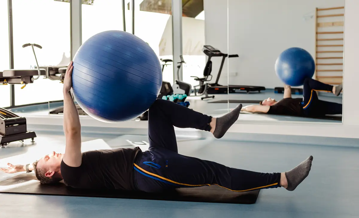 Pacient întins pe saltea neagră execută exercițiu de stabilitate de trunchi cu minge fitness albastră între mâini și genunchi; sală luminoasă cu bandă și echipamente Motion Clinic, Snagov Plaza.