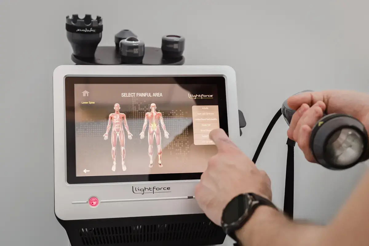 Consolă laser terapeutic LightForce XPi cu ecranul „SELECT PAINFUL AREA”, aplicator în mână și zona lombară evidențiată pe anatomie — echipament Motion Clinic, Snagov Plaza.