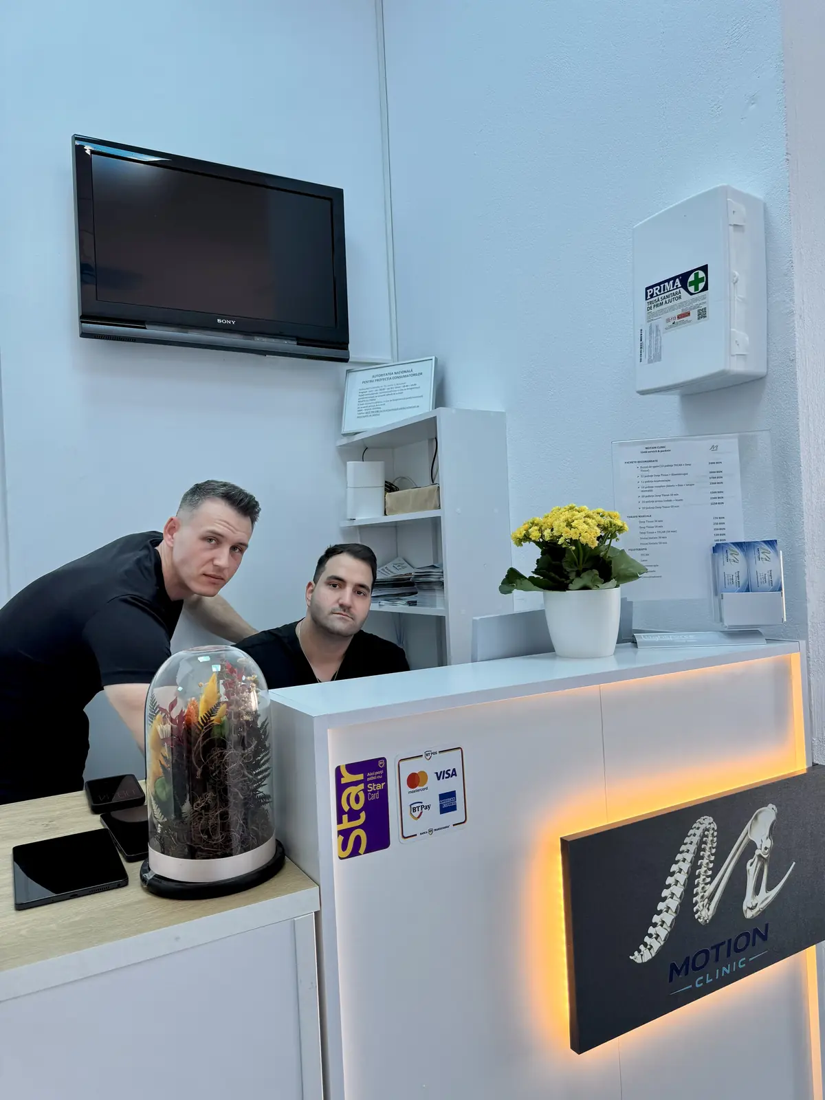 Recepția Motion Clinic — două persoane în spatele tejghelei albe, logo iluminat Motion Clinic și stickers plată card; perete alb, trusă de prim ajutor — Snagov Plaza, Ilfov.