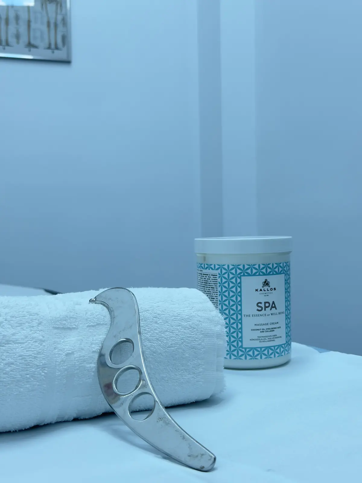 Unealtă IASTM, prosop și cremă Kallos pe fundal bleu; colț poster anatomic — detaliu pregătire masaj și mobilizare țesuturi, Motion Clinic.