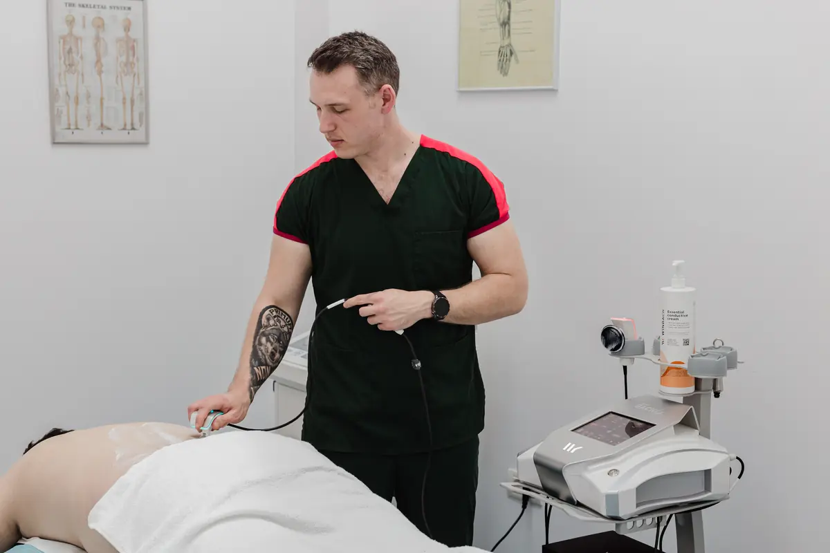 Terapeut aplică aplicator Winback cu gel pe spatele pacientului în prona; consolă TECAR și postere anatomice — plan combinat terapie manuală și fizioterapie, Motion Clinic.