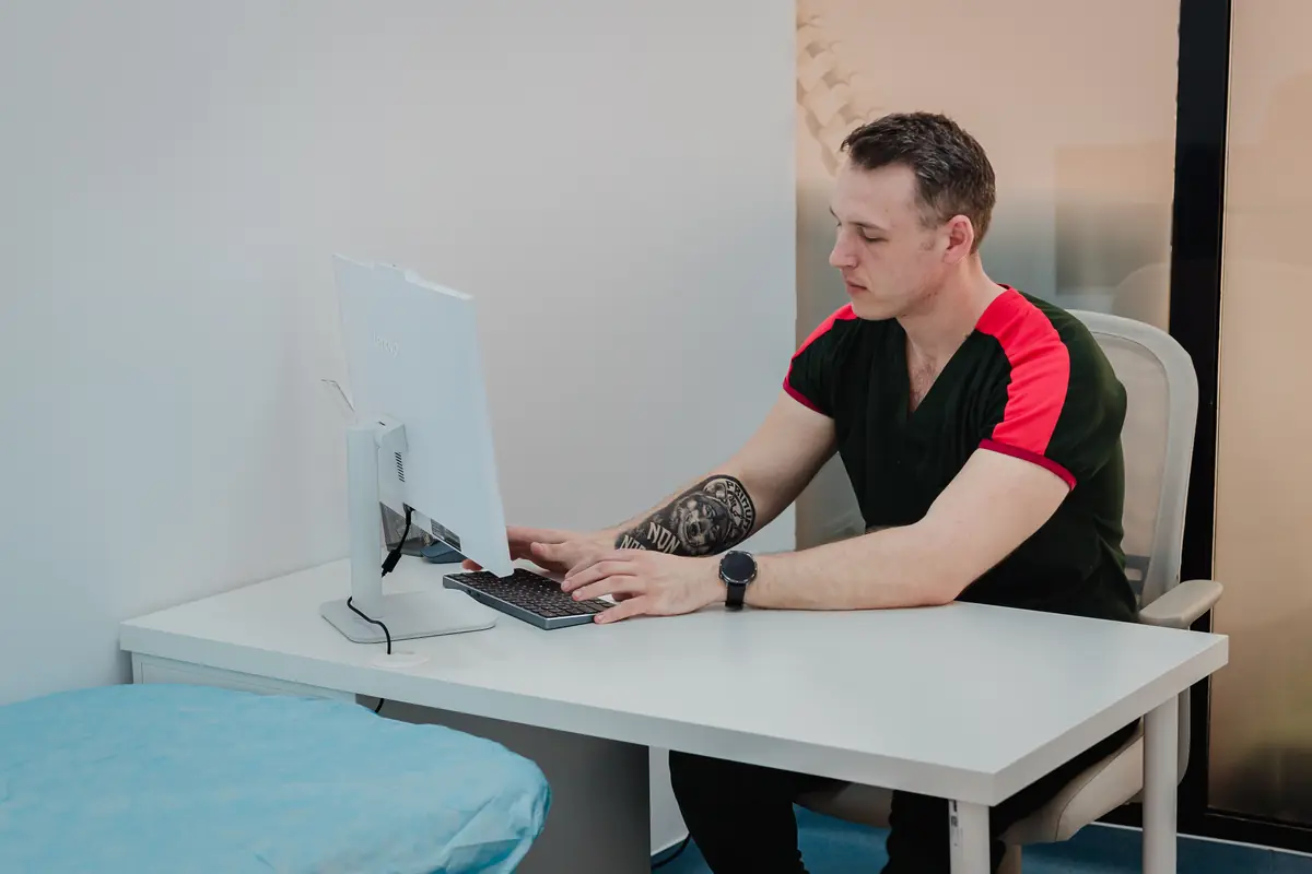 Specialist Motion Clinic la birou cu monitor; în prim-plan colț masă de tratament acoperită — planificare și documentare în clinică, Snagov Plaza.